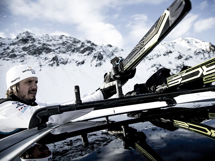 A close-up of a man parked in the snow loading skis into a ski rack for car.