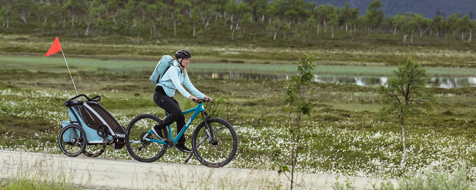 Женщина едет по полю на велосипеде с синим велоприцепом Thule Chariot Cross.