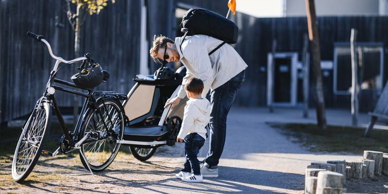 A father leans down to his toddler who is standing beside a bike and a Thule bike trailer for kids.