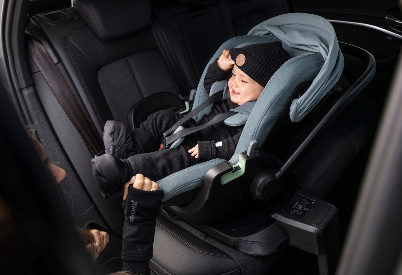 A baby smiling in a Thule Maple infant car seat mounted on the base. The seat is turned outwards and a person’s hands are touching the edge of the car seat.
