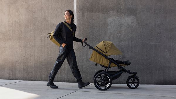 A woman walks down a concrete street with a Nutria Thule Urban Glide 3 all-terrain stroller.