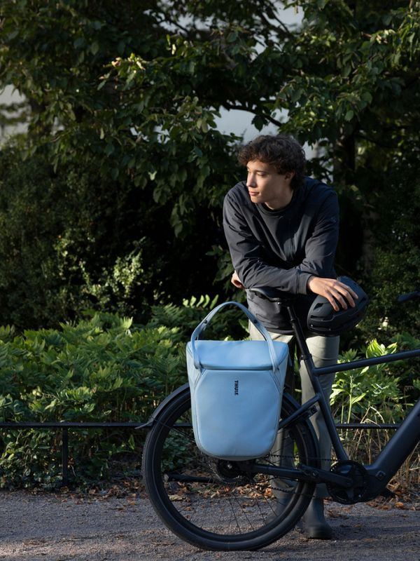 Un homme, un vélo noir et un sac bleu devant des arbres verts.