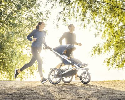 Een man en vrouw joggen in de zon met hun kind in een sportieve kinderwagen.
