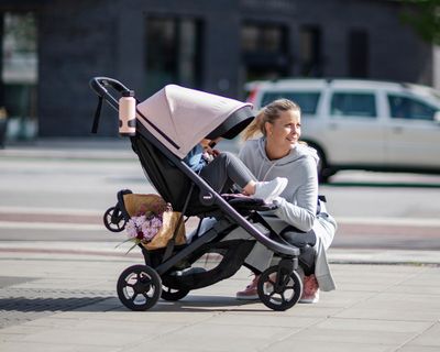 A woman sits next to a pink stroller with various stroller accessories.