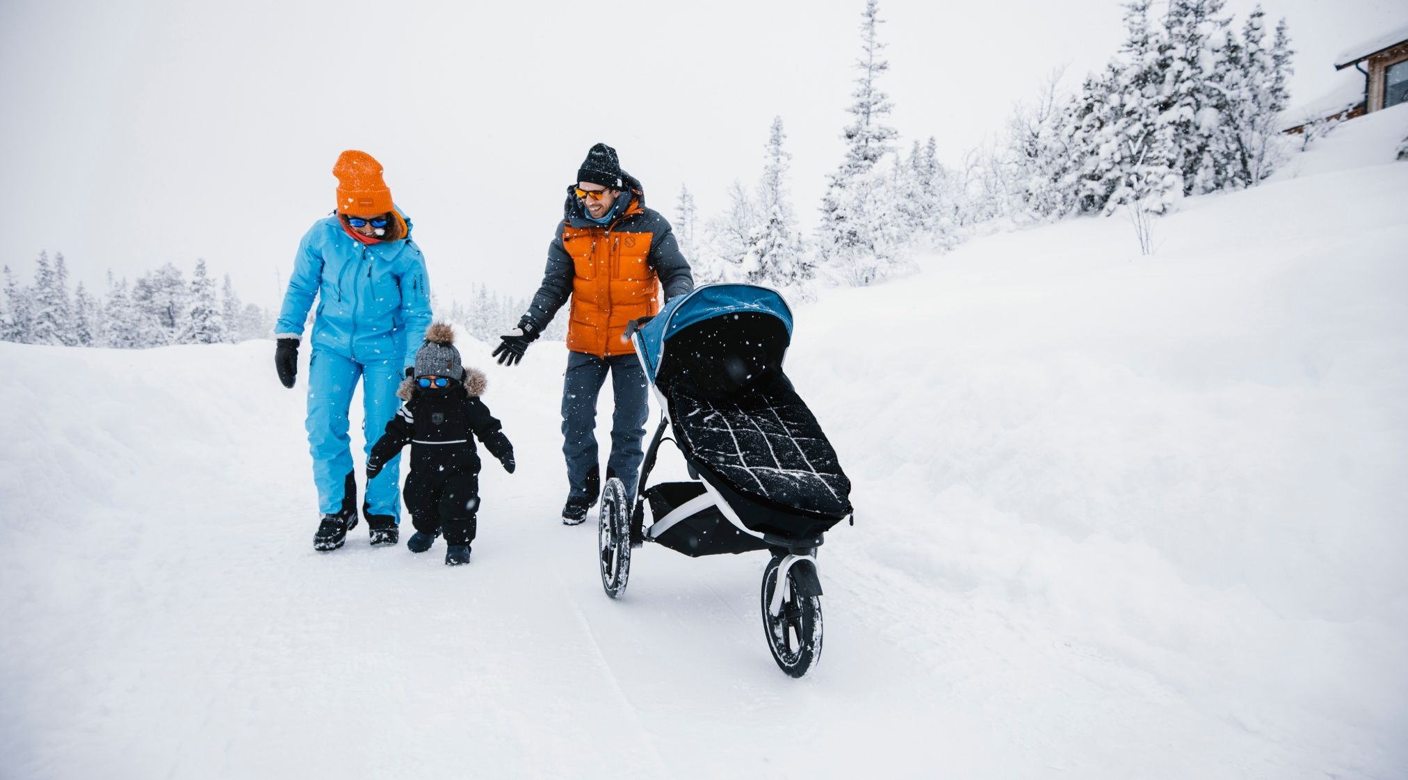 Two parents walk through the snow with their child in a snow suit and a stroller with the best stroller accessories for the snow.