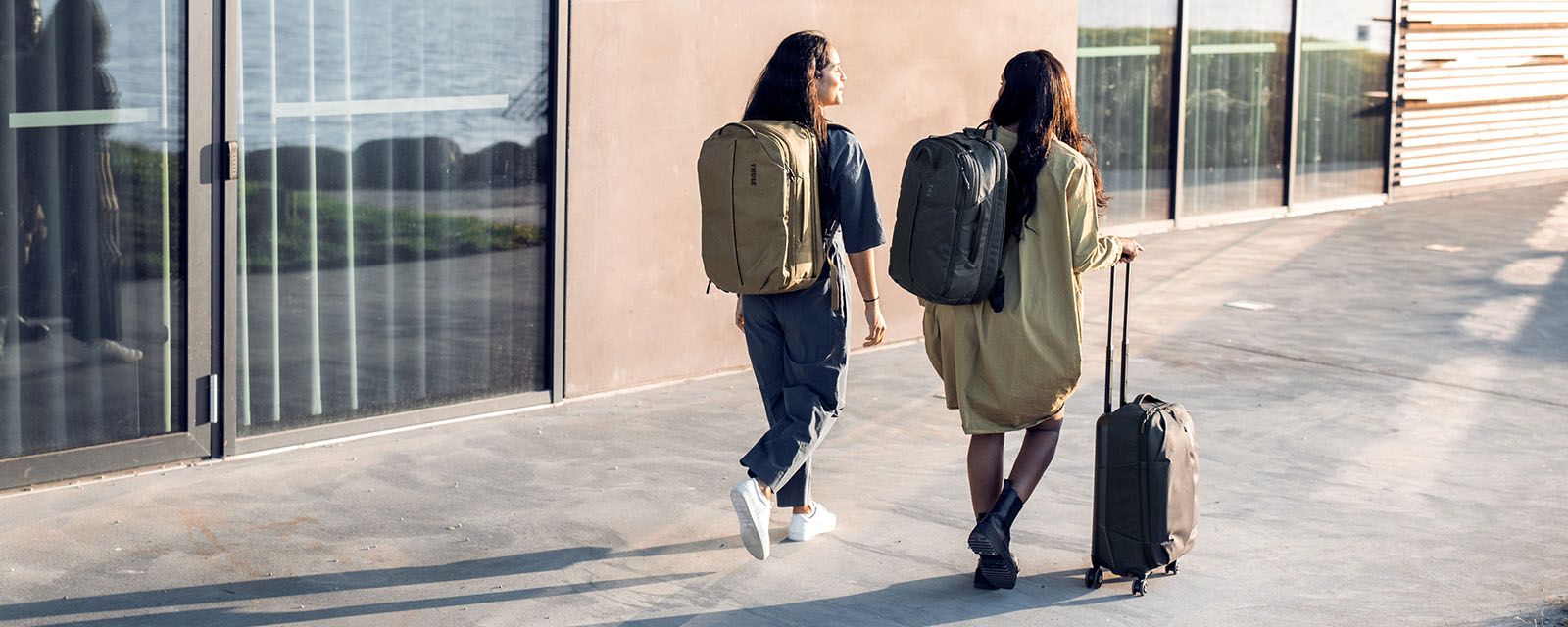 Zwei Frauen mit Thule Aion Rucksäcken und einem Handgepäckkoffer gehen einen sonnigen Gehweg entlang.