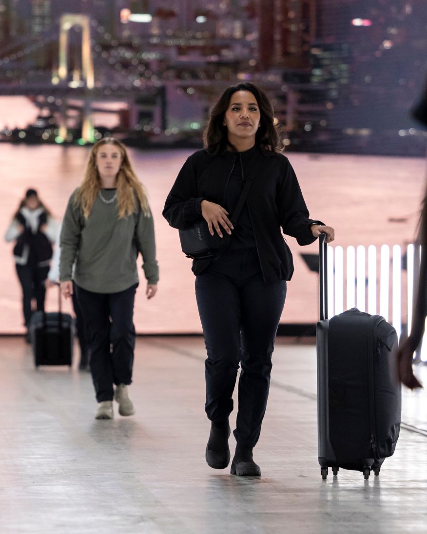 A medium dark-skinned woman in all black clothes walking down a fashion runway rolling a Thule Subterra 2 suitcase and wearing a small crossbody bag. Behind her is a woman with long blonde hair and another blurred person walking  down the same runway.
