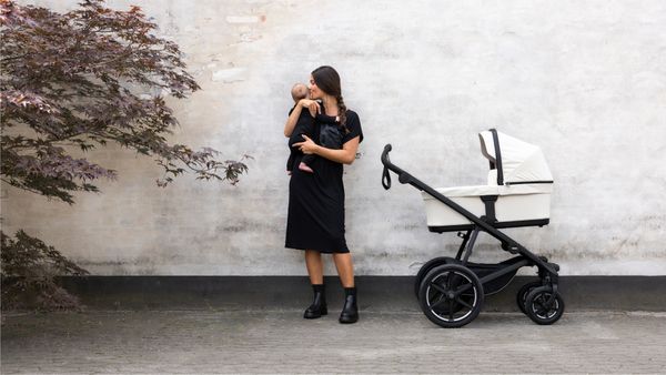 A woman stands against a while wall holding her baby next to a white bassinet Thule Urban Glide 4-wheel stroller.