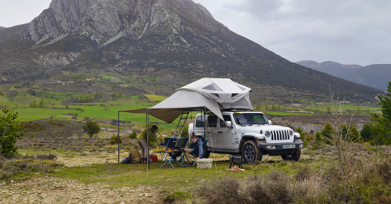 Two people sit in camping chairs under the awning of a jeep with a Thule Approach rooftop tent that is parked in the mountains.
