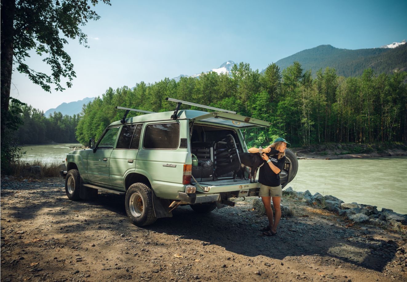 Uma pessoa está de pé junto a um automóvel estacionado na margem de um rio, com a bagageira aberta. A pessoa faz festas a um cão sentado na bagageira do automóvel em frente a um transportador para cães Thule Allax aberto.