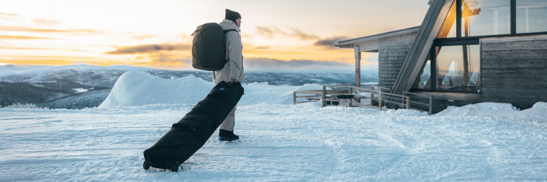 Uma pessoa a caminhar por uma paisagem nevada carregando uma grande mala e puxando um esqui ou uma mala de snowboard em direção a uma cabana ao pôr do sol.