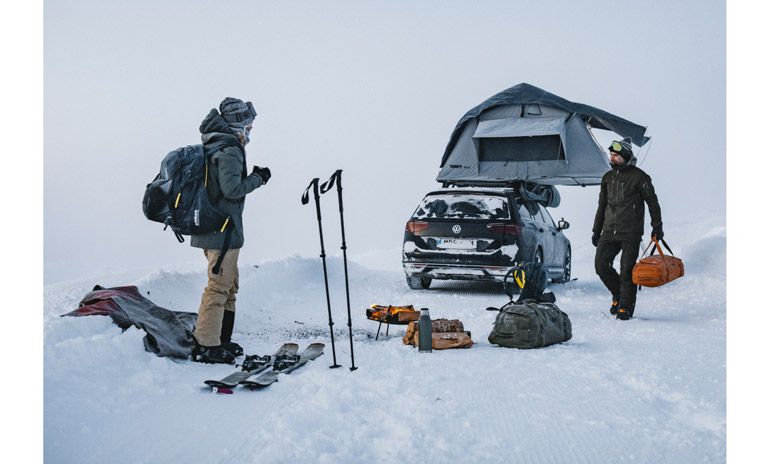 Two men with gear stand in a snowy landscape with a car parked and a Thule Tepui rootop tent on the roof.