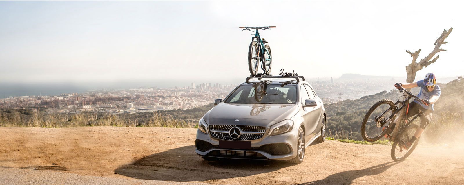 Mountain biker on a bike next to a car with a Thule roof bike rack with a city in the background.