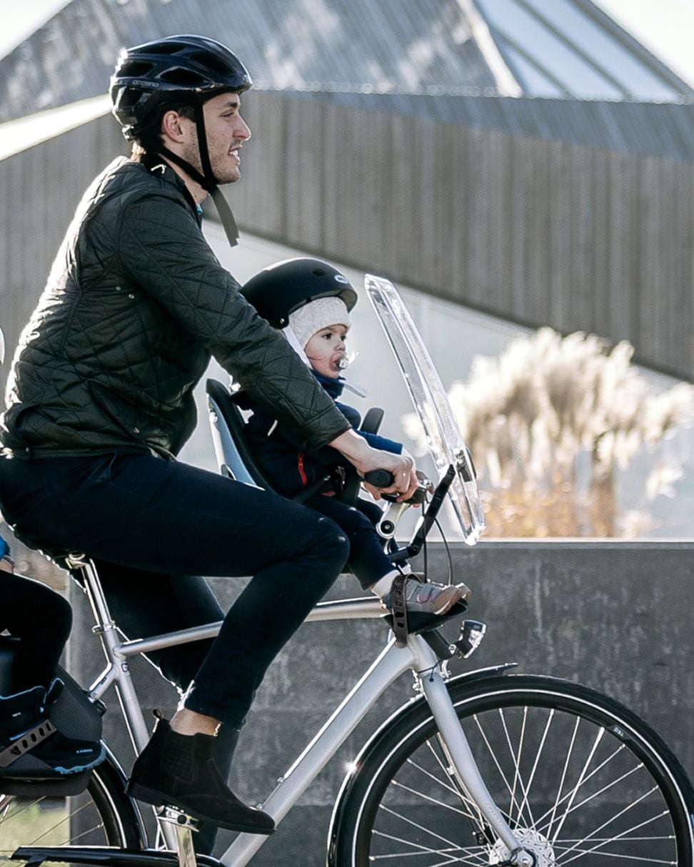 Adult riding a bicycle with a front-mounted child seat fitted with a wind shield accessory, carrying a small child, traveling along a paved urban path.