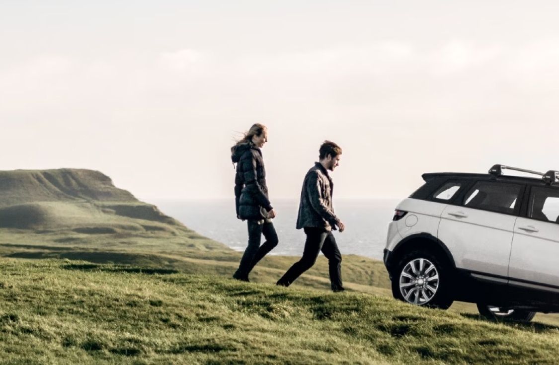 두 사람이 바다 근처의 탁 트인 풍경 속에서 Thule 루프 랙이 장착된 차를 향해 걸어갑니다.