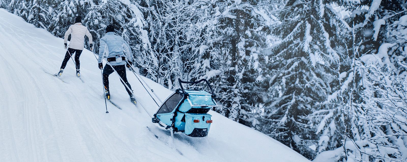 Een stel gaat langlaufen op een besneeuwde heuvel met hun kind in een Thule Chariot Cross skitrailer