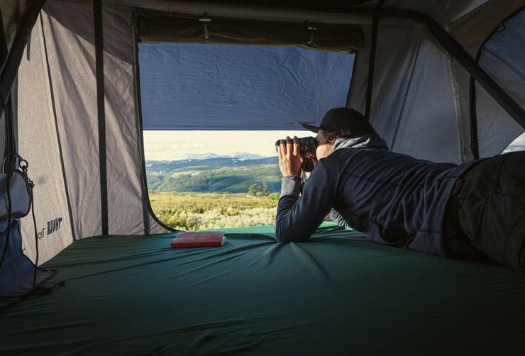 A man inside a Thule Tepui roof top tent looks out at the mountains with his binoculars.