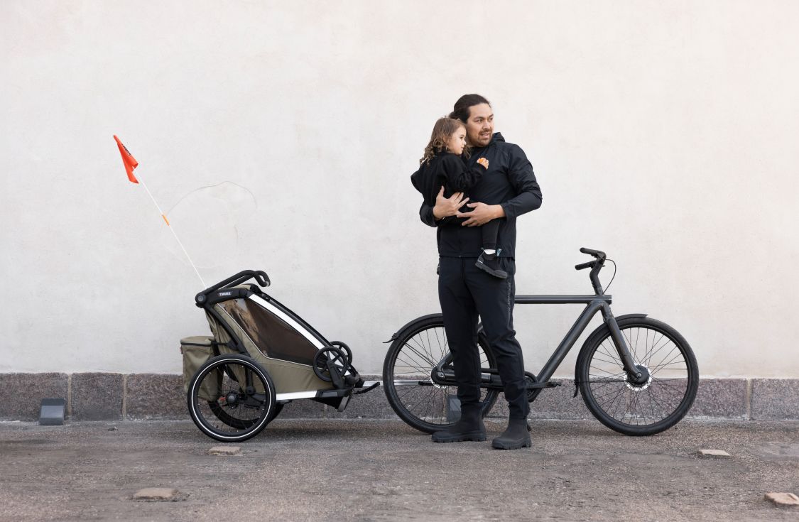 Мужчина с ребенком на руках стоит перед мультиспортивным велосипедным прицепом Thule Chariot, который прикреплен к велосипеду.