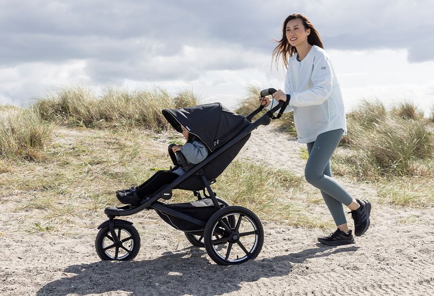 A woman is pushing a Thule Urban Glide 3 stroller with a child in it across the sand.