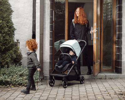 En kvinna går ut genom en trång dörröppning med en kompakt barnvagn och barnet springer framför.