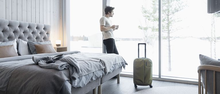 Um homem num quarto de hotel olha pela janela com um café na mão e uma mala ao lado.