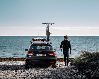 Eine Person geht vom Auto aus dem Meer, das auf einem kieseligen Ufer geparkt ist und auf dem Deck ein Surfbrett montiert ist.