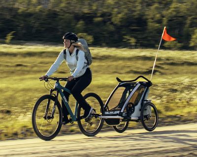 Une femme fait du vélo dans un champ avec une remorque pour vélos Thule Chariot bleue.