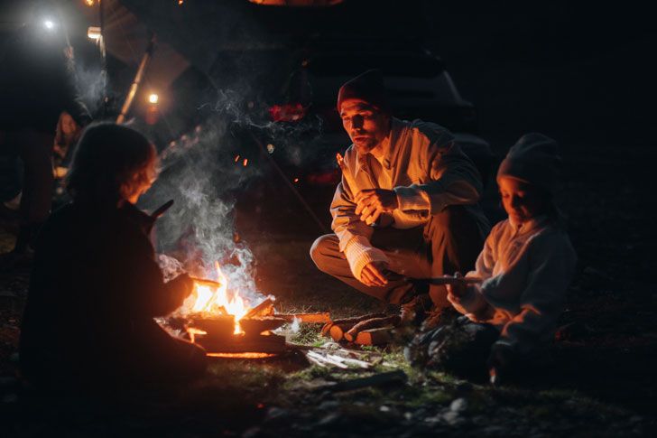 Een gezin met kinderen zit aan een kampvuur in het donker naast een Thule daktent.