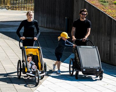 Due genitori passeggiano nel sole lungo una via cittadina trasportando rimorchi da bici per bambini.
