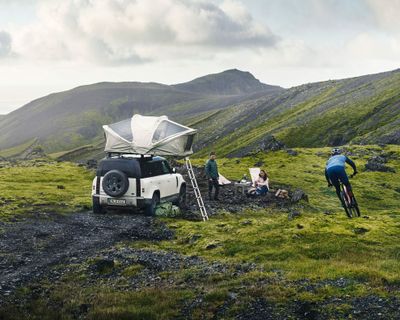 A person bikes next to a campsite where an SUV is parked with a Thule rooftop tent on top