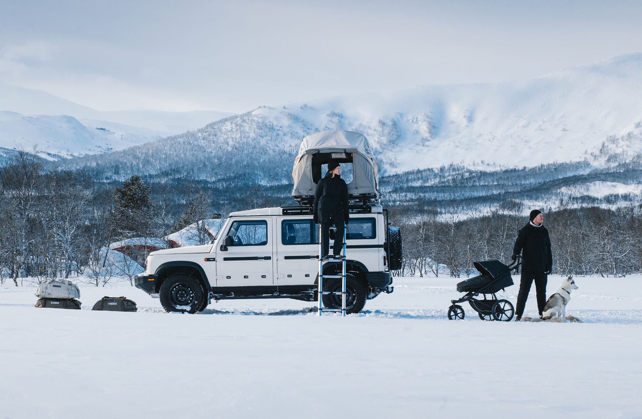 A winter camping scene with a white off-road vehicle equipped with gear from Thule, including a rooftop tent and other outdoor essentials, surrounded by snowy mountains.