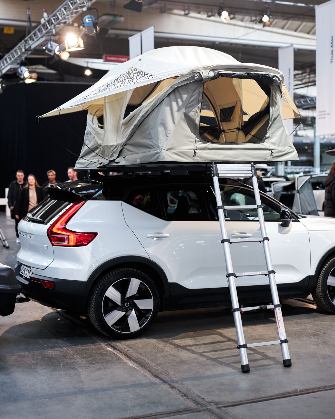 A white Volvo XC40 with an unfolded Thule Approach rooftop tent parked in a large exhibition hall. There are metal stairs leading to the open rooftop tent and people standing behind the car.