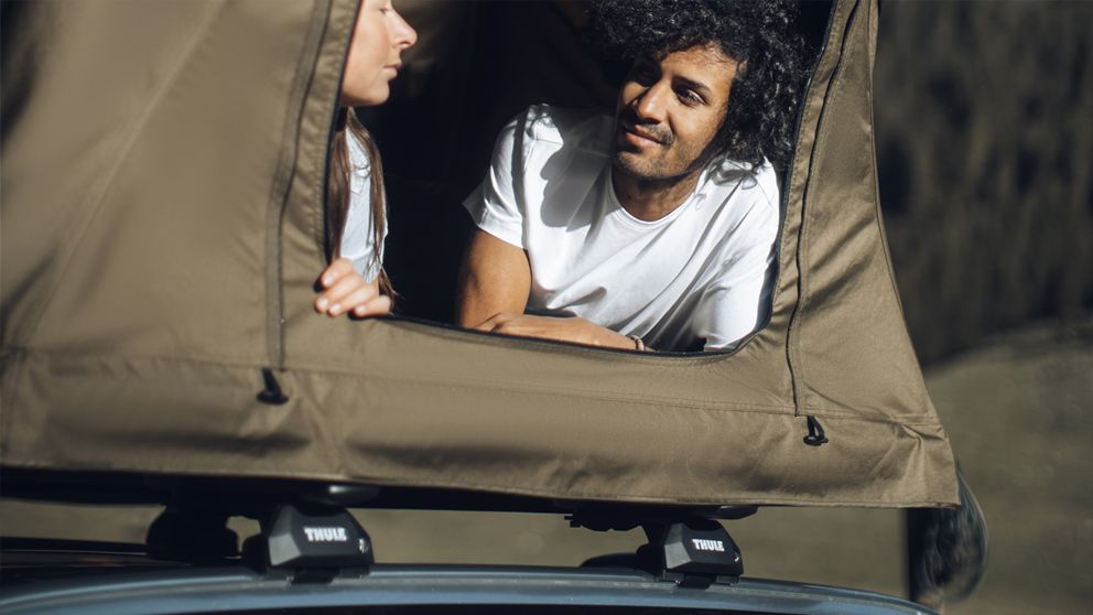 A man and woman sit in a roof top tent on Thule roof racks looking at each other.