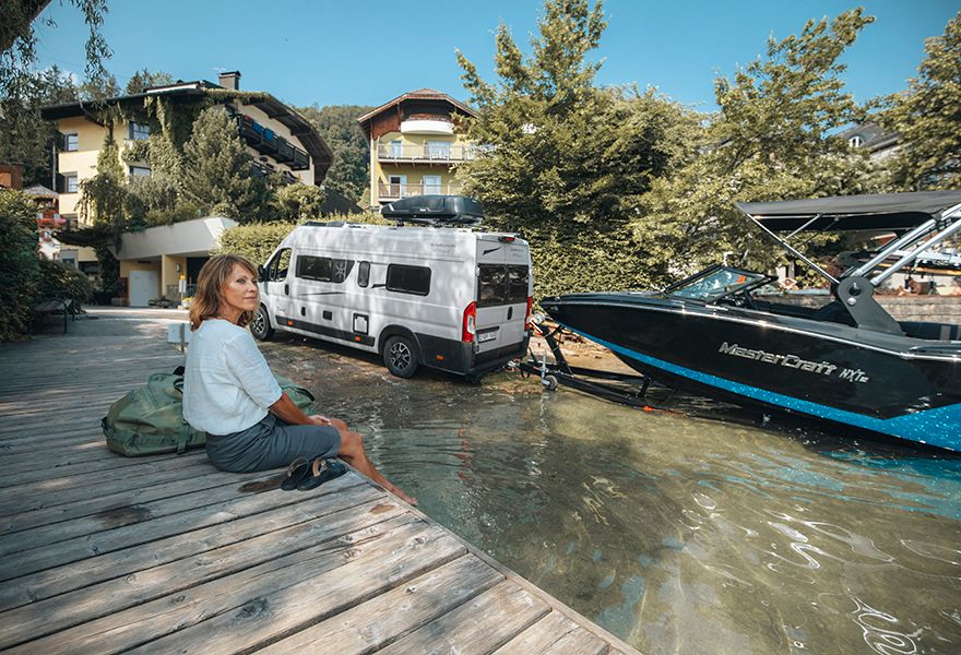 By the water, a woman watches a white van tow a boat thanks to the Thule VeloSwing towbar.