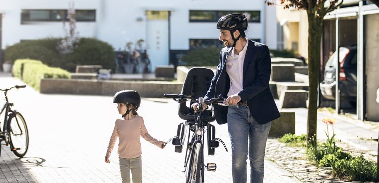 A man walks with his daughter holding a bike with a kids bike seat.