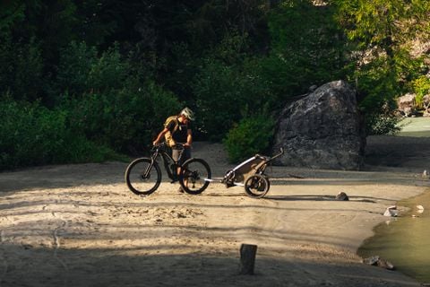 Person standing with a bicycle, pulling a bike trailer along a sandy riverbank.