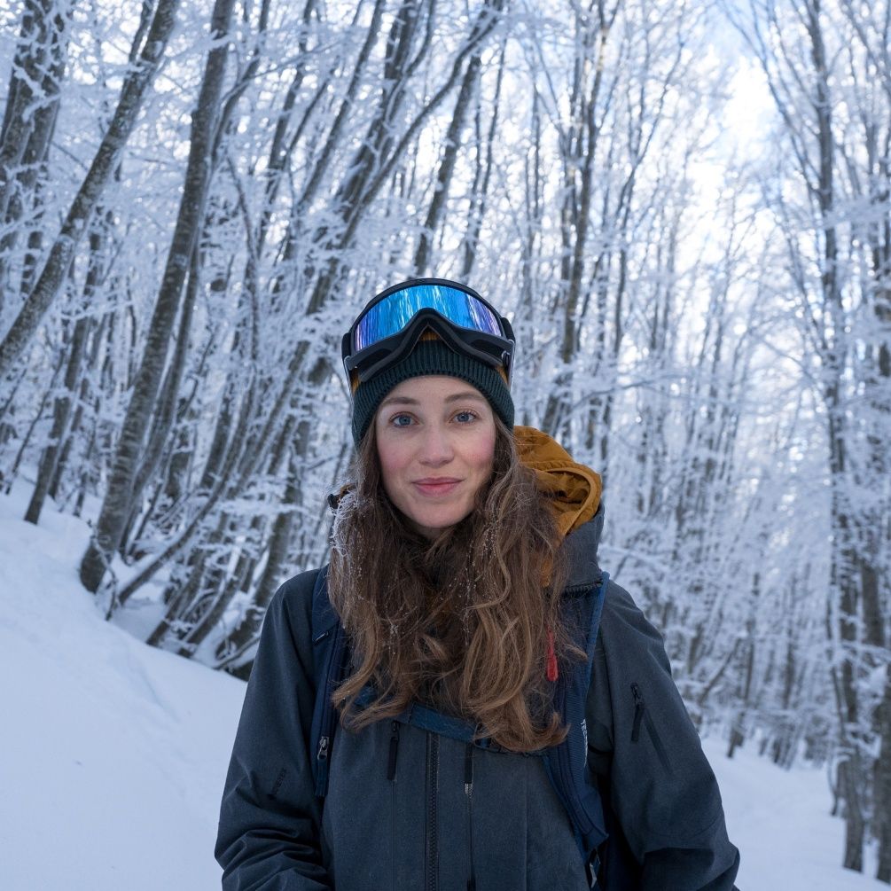 A close up image of Alice Linari smiling at the camera in a snowy forest with snow goggles on her head.