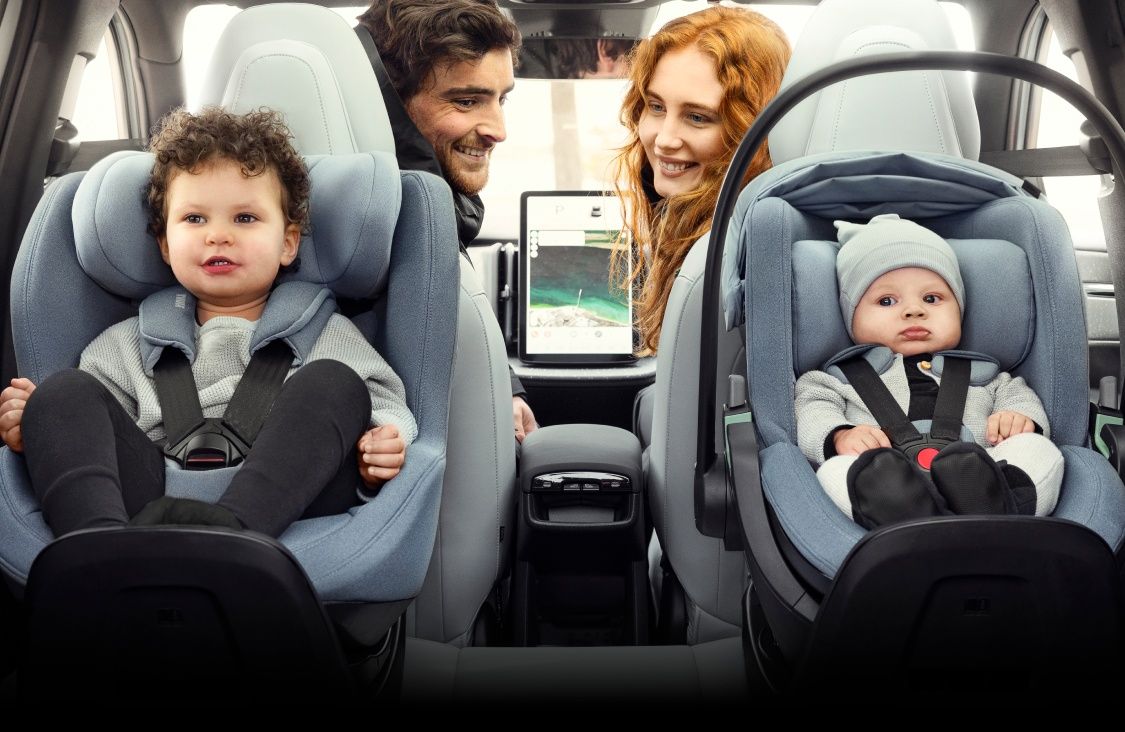 Two children sit in rear facing car seats while two smiling adults look back at them from the front seats of a vehicle.