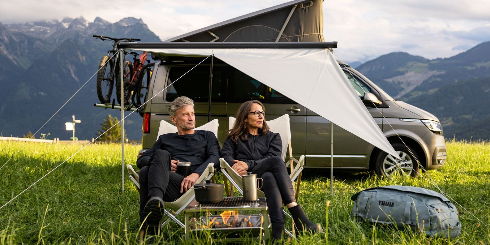 A man and woman sit in camping chairs in front of their compact van with a Thule Subsola awning panel.