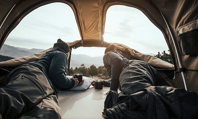 Kaksi ihmistä makaa makuupusseissa Thule Approach ‑kattoteltassa ja katsoo panoraamaikkunoista avautuvaa maisemaa. 