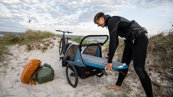 一位穿着潜水服的男士在沙滩边将冲浪板装进儿童载物自行车拖车中。