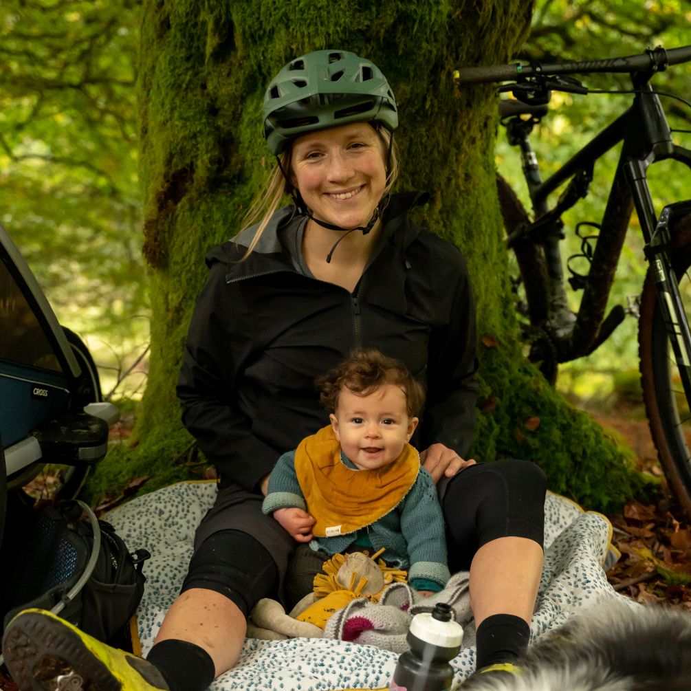 A woman with her helmet and her baby sitting on a blanket and leaning towards a tree.
