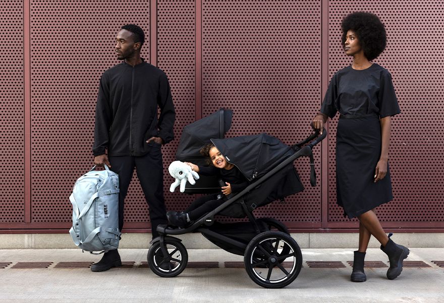 A man is holding a Thule duffel bag and a woman is standing next to the Thule Urban Glide 3 double stroller with a smiling child in it.
