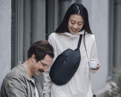 A woman holding a coffee and wearing a tote bag smiles at a man looking down at a notebook.