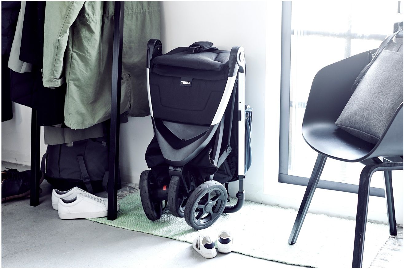 A stroller is folded up compactly and placed in the corner of a room next to a hanger.