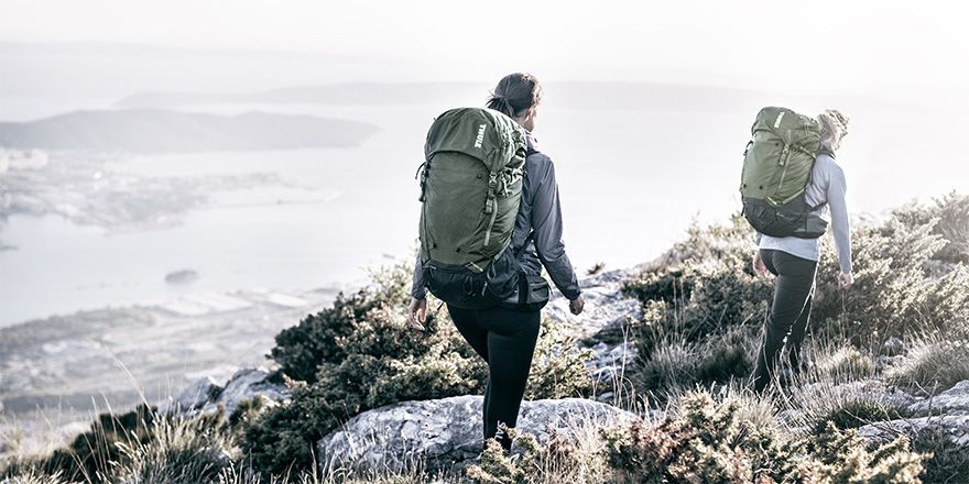 Two people walk along a mountain with a view of the ocean and a Thule hiking backpack.