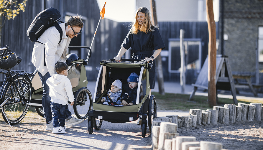 Famille avec un vélo et deux remorques pour enfants devant une aire de jeux.