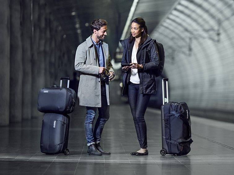 A man and a woman stand waiting in a subway station with their Thule checked luggage.