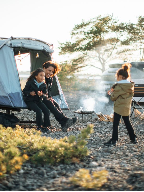 Une mère et deux enfants sont assis à l'entrée d'une tente Thule Outset montée sur attelage, profitant d'un moment chaleureux dans un cadre extérieur sauvage. Un feu de camp crépite à proximité, tandis qu'un autre enfant réchauffe ses mains.
