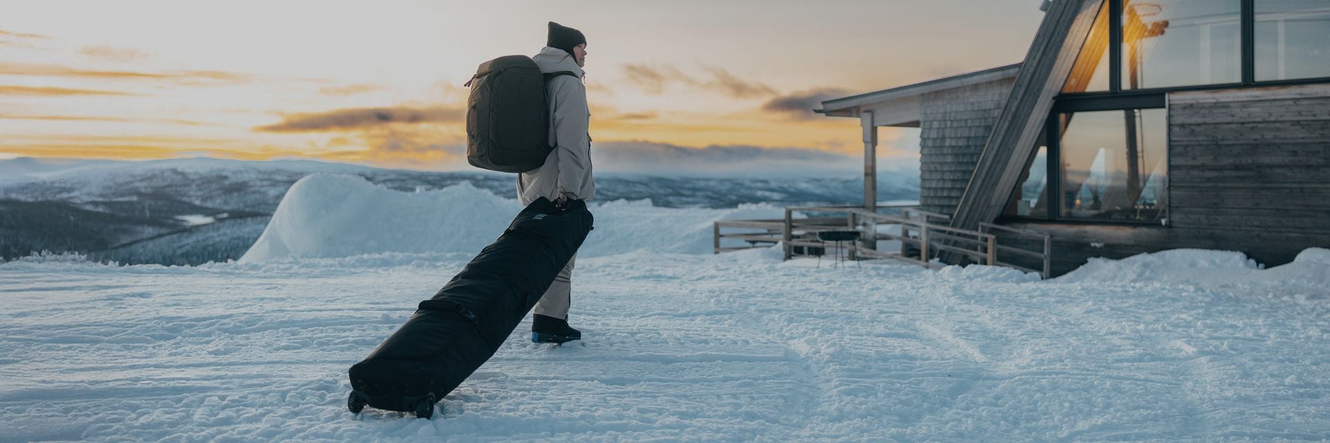 Person carrying Thule ski bags through a snowy mountain landscape at sunset.